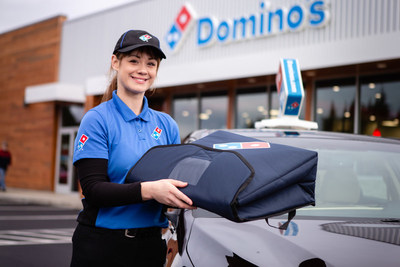 Chauffeuse-livreuse debout  ct de sa voiture devant une succursale de Domino's Pizza (Groupe CNW/Domino's Pizza)