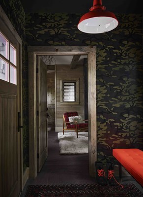 A hunting blaze orange door welcomes guests into a foyer featuring a bench of the same color accented by custom camouflage wallpaper. The space offers a peek through to the living room beyond.