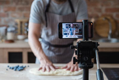 baker online courses. food preparing and culinary training class concept. smiling bearded chef kneading dough in the kitchen and shooting video of himself using mobile phone on a tripod. (CNW Group/Thinkific Labs Inc.)