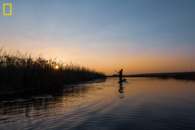 A poler navigates on the Cuando River at sunset. The Okavango Wilderness Project's 2018 expedition focused on the eastern-most section of their survey area in Angola. The trek took the team down the length of the Cuando River, a journey that allowed them to explore the intersection of the Okavango and Zambezi Basins, two of the largest basins in southern Africa. Photo by Kostadin Luchansky / National Geographic Okavango Wilderness Project.