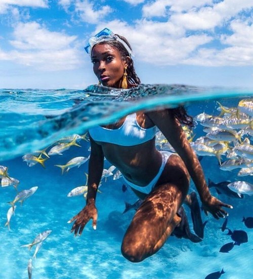 Young black woman snorkeling in the ocean wearing goggles, fins, a white two-piece bathing suit, surrounded by exotic fish