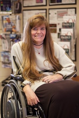 Image description: Kim Anderson-Erisman, PhD, a white woman with long, light brown hair, sits strapped into her manual wheelchair, wearing a light shirt with striped long-sleeves and documents hanging on the wall behind her.