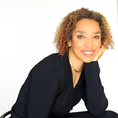 Image description: Alice Sheppard, a multiracial Black woman with coffee-colored skin and short curly brown and golden hair, leans forward resting her cheek on her hand. She wears a black shirt and a gold necklace and smiles at the viewer. Photo by Beverlie Lord.