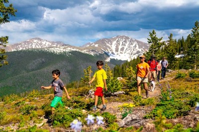 Sunny skies, fresh mountain air surround a family hiking at Winter Park Resort in Colorado. The Continental Divide and 13,397-foot Parry Peak rise up behind them.