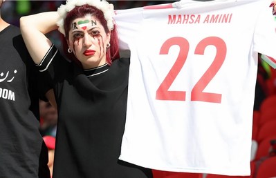 Iran Soccer Team Begrudgingly Sings Anthem Then Plays Their Hearts Out-An Iranian protestor at the World Cup holds a jersey with the name Mahsa Amini, who recently died under police custody at the age 22.