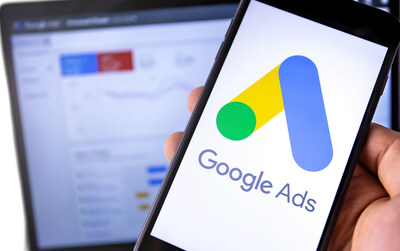 A hand holding a phone that has Google Ads on the screen with a green dot, and yellow and blue lines. There is a computer screen with a line graph in the background. (CNW Group/Unifor)