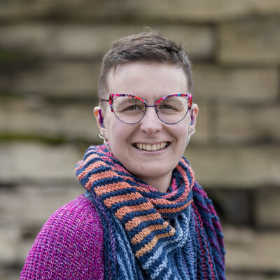 Angela Adler is a white nonbinary disabled and neurodivergent millennial with olive skin and green eyes, short brown hair, purple hearing aids, and pink and purple cateye glasses, wearing a magenta sweater, a blue and jewel-tone striped scarf, and silver earrings, pictured smiling in front of a weathered wood wall.