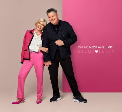 Selma Blair, QVC's Brand Ambassador for Accessibility, stands hand in hand with award-winning fashion designer Isaac Mizrahi, smiling on a photo shoot set. Selma wears a pink blazer with a white blouse, a pearl necklace, pink pants, and heels. Issac is dressed in dark colors. The background is a color block split screen of neutral and pink.