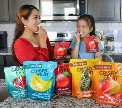 Mother and daughter snacking on Crispy Fruit Apple, with bags of Crispy Fruit Strawberry, Banana, Apple, Pineapple and Mango in front of them on the kitchen counter.