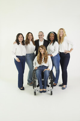 Five diverse models are showcasing NYDJ's Marilyn Straight Adapt-Denimtm jeans alongside Brand Ambassador Mark Peters. All models pose in various denim hues with white tops, while Mark, donning a white shirt and black sportscoat, stands among them. In the forefront, a wheelchair model poses elegantly.
