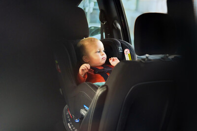 An infant secured in a car seat is alone inside a vehicle, looking out of the window. The car seat is black, and the child is wearing a red outfit. The interior of the car is dark-colored.