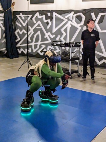 An individual wearing EKTO VR's robotic footwear and a VR headset, squatting during a VR session, with an EKTO VR team member observing in the background.