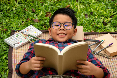 Asian boy in red shirt having fun reading book under tree in park