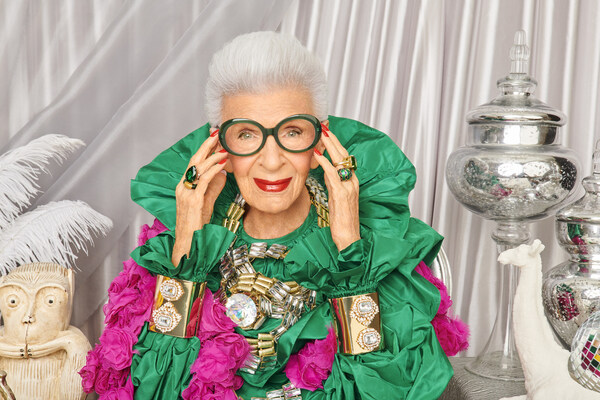 Woman wearing oversized green ruffled dress, large round glasses, and colorful jewelry.