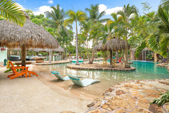 The resort's stunning pool area features a sandy zero-entry, four tiki huts, an outdoor kitchen, a bar, and lush tropical landscaping, creating a relaxing and private oasis for guests.