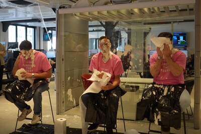 Three individuals wearing pink 'Funding the Fight' shirts sit on stools with whipped cream pies on their faces, smiling and wiping off the mess in a bright office setting, showing their support for breast cancer awareness.