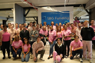 Group photo of SBG Funding employees wearing pink and standing together in the office in support of Breast Cancer Awareness Month, with many team members smiling in front of the company logo.