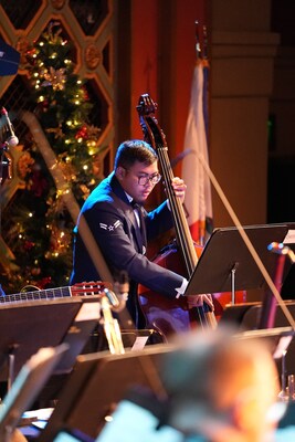 Close up on a musician in the Air Force Band of the Golden West playing an upright bass with the concert band, with a festive tree and holiday decorations in the background.