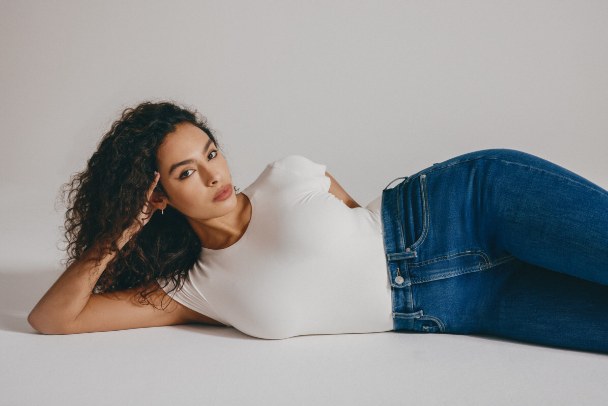 Modell i vit topp och blå Spanx ReDefine-jeans, liggande mot en ljus bakgrund.