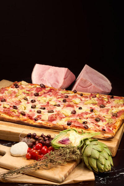 Angelina Bakery’s gourmet pinsa layered with ham, artichokes, black olives, and mozzarella—served with rustic Mediterranean ingredients.