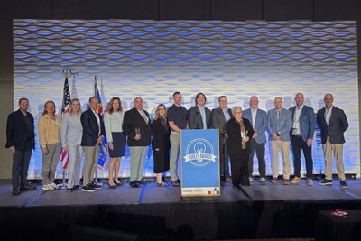 A group of sixteen people stands on a stage in front of flags.