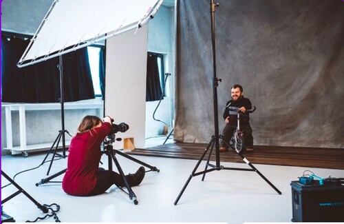 Image of a photo shoot with two people of short stature. One is wearing a red shirt and black pants taking an image of another Little Person is posing for the camera on scooter and smiling.