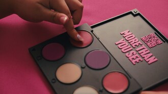 A close-up of a hand selecting a reddish shade from a colourful makeup palette. The palette lid features raised pink text that reads &#8220;MORE THAN YOU SEE&#8221; alongside a QR code, set against a bright pink background. (CNW Group/Shine Foundation)