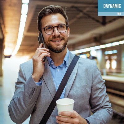 A man wearing glasses and a business suit talks on a mobile phone while holding a paper coffee cup, standing in a brightly lit indoor space that appears to be a transit station. The image features a blue-and-white box in the upper-right corner with the text "DAYNAMIC PRO".