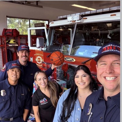Group photo inside a fire station showing Rooter Hero representatives Alexandria Lavoie (center left) and Katherine Conchas (center right) smiling alongside Fullerton Fire Department firefighters in front of a fire engine, as they deliver donated toys for the Rooter Hero toy drive.