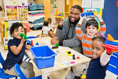 An adult and three small children smile at the camera while holding up what they have built with blocks while sitting at a table in a preschool classroom.
