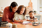 A caregiver smiles while helping a young child write in a notebook at a kitchen table, as another child sits nearby with a laptop and headphones.