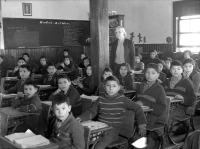 Cree students at their desks with their teacher in a classroom, All Saints Indian Residential School, Lac La Ronge, Saskatchewan, March 1945 (CNW Group/Royal Canadian Geographical Society)